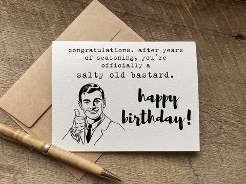 A novelty birthday card with a black-and-white illustration of a man giving a thumbs-up that reads: "congratulations. after years of seasoning, you’re officially a salty old bastard. happy birthday"