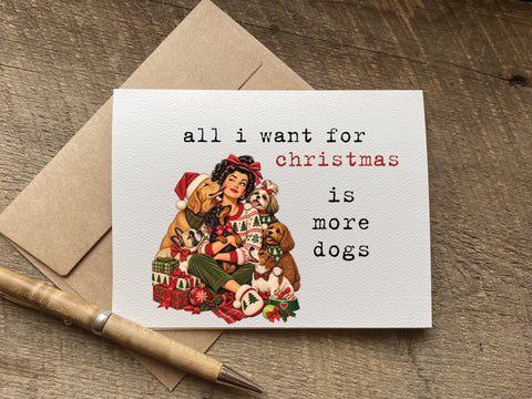 A funny christmas greeting card with a vintage-style illustration of a woman and three dogs wrapped in holiday clothing, The card reads, 'all i want for christmas is more dogs' in black and red text.