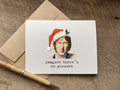 A white greeting card with a humorous illustration of John Lennon wearing a Santa hat and yellow glasses, with the text 'imagine there's no present'. 
