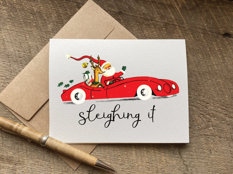 Christmas card with vintage graphic of Santa Claus driving a red car and the text 'sleighing it' 